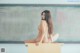 A naked woman sitting on top of a desk in front of a blackboard.